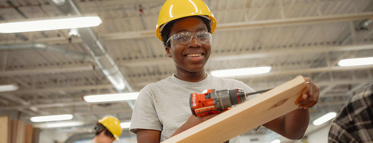 A smiling kid in hard hat and glasses works on a piece of wood at an NSCC trade camp.