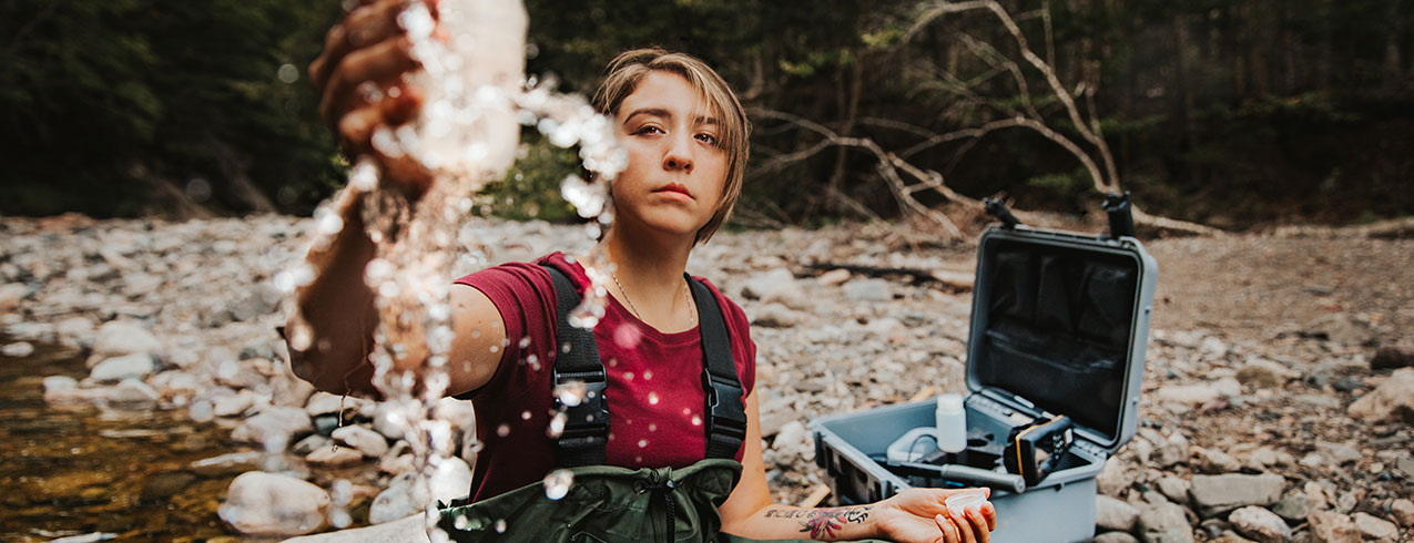 A female student is seen sitting in a river wearing protective coveralls and boots with a large case of instruments beside her for testing the water.