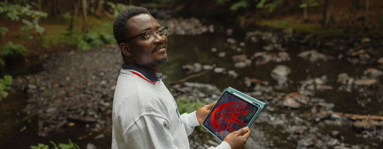 A person standing in a wooded area, holding an iPad with a wildfire map on its screen. 