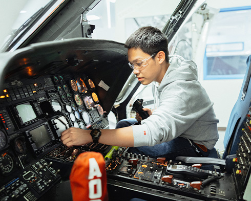 Person seated in an aircraft cockpit, reaching toward illuminated control panels and flight instruments during a hands-on training or maintenance session.
