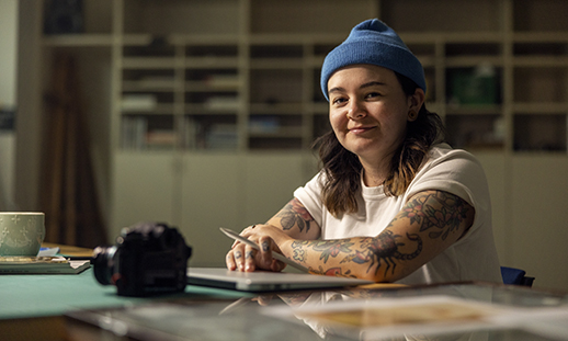 A person wearing a beanie hat and t-shirt with tattooed arms sits at a table in front of a closed laptop. She looks at the camera with a slight smile.