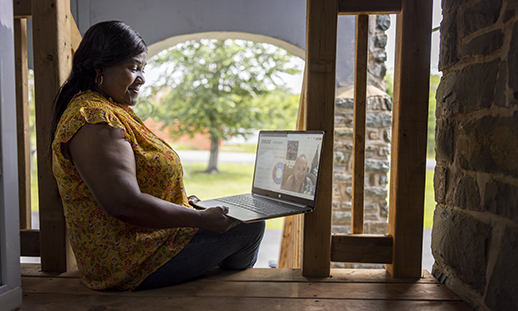 A woman sits on her front steps and holds an open laptop on her lap while taking part in online classes.