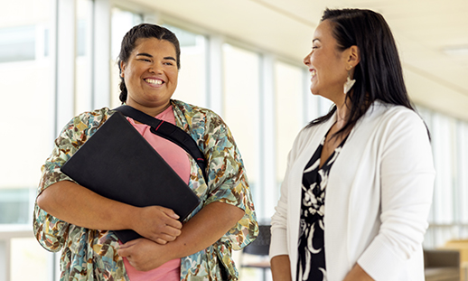 A woman wearing a t-shirt and scarf holds a textbook against her chest and smiles and she has a conversation with an NSCC student advisor.