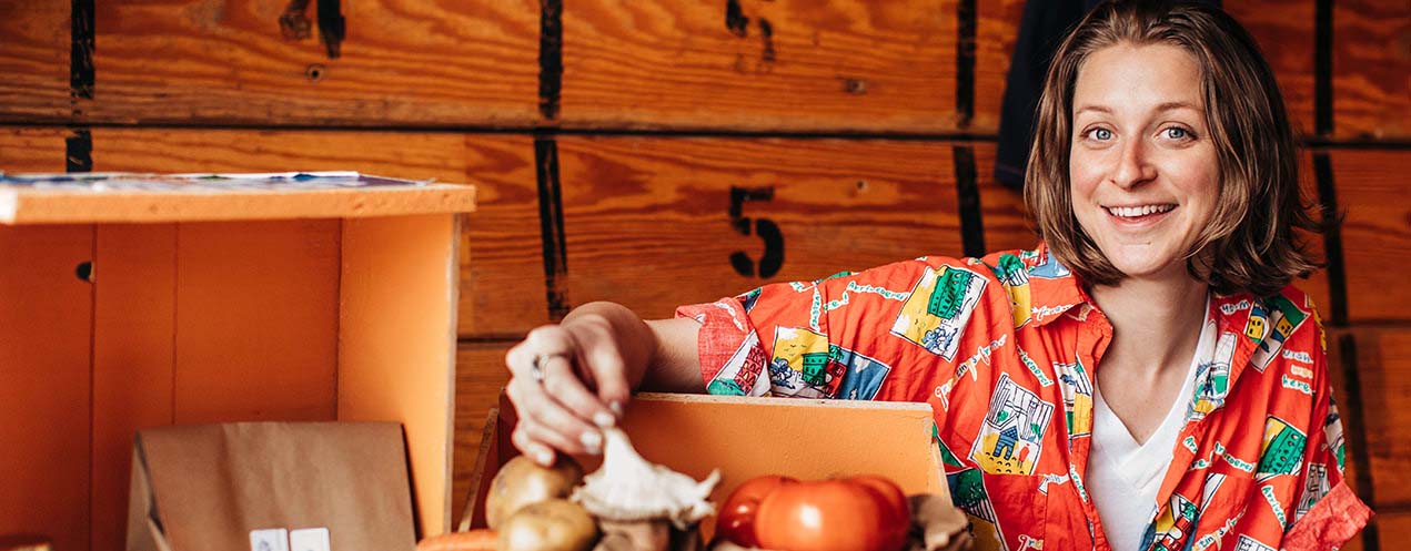 Kara Friesen, an NSCC graduate, sits in front of a box of produce and holds a mushroom while smiling at the camera.