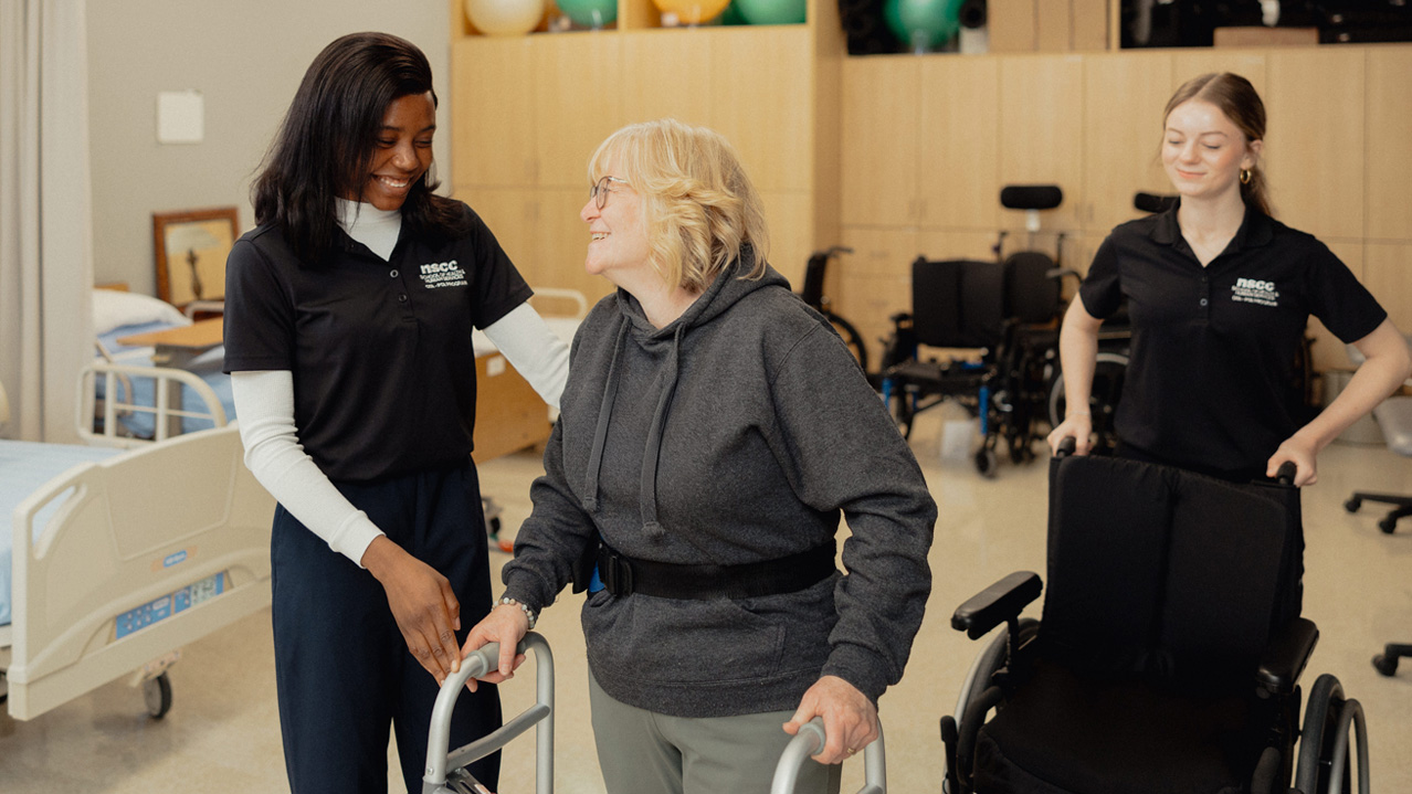 Two students assist a patient using a walker. All 3 smile while interacting in a hospital-like setting.