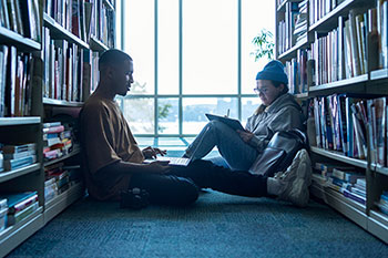 Two people sit on the floor between shelves in a library with open laptops.