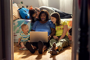 A woman sits on the floor with an open laptop with 2 children sitting on either side of her and an adult on the bed behind her.
