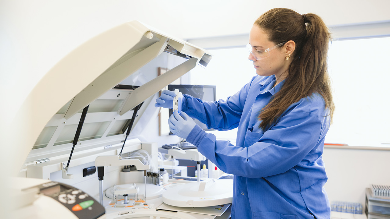 A medical lab technologist in a lab coat wears protective glasses and gloves while holding 2 vials in a laboratory.