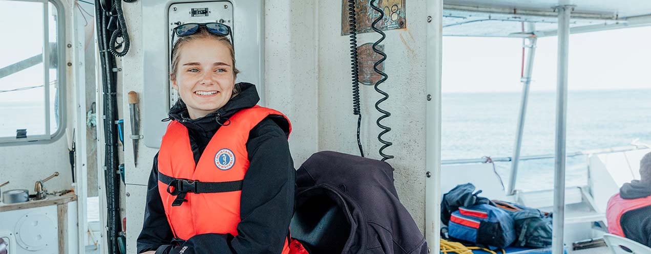 A person sits on a boat at sea. She wears a jacket and personal floatation device. Her hair is pulled back and sunglasses rest on the top of her head. She smiles while looking slightly to her left.