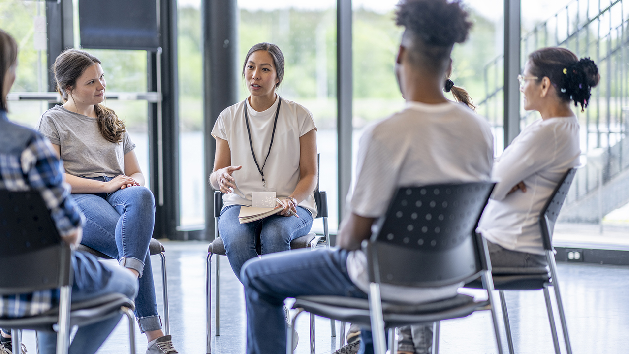 A group of youth and an adult sit in a circle in chairs and have a discussion led by the adult.