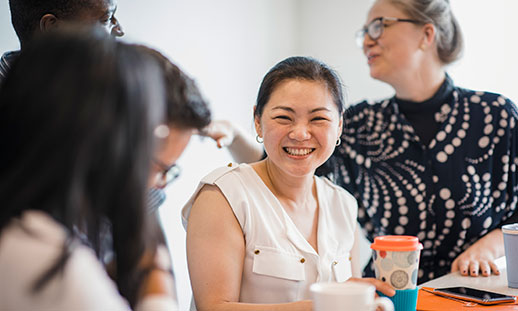 A group of 5 employees sit at a table. Several employees are engaged in conversation. One employee looks at the camera and smiles while holding a coffee mug.