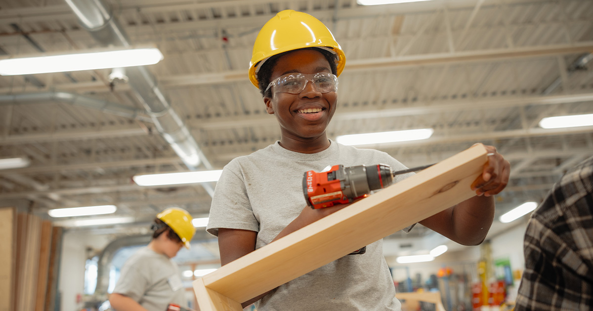 A student holding a carpentry project and a power tool, smiling and looking at the camera