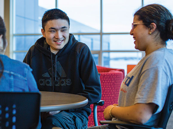 Three people talk and laugh while sitting at a round table in a library.