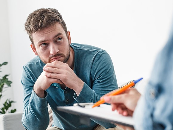 A man sits with his chin resting on his hands and looks left toward a person holding a clipboard and pen.