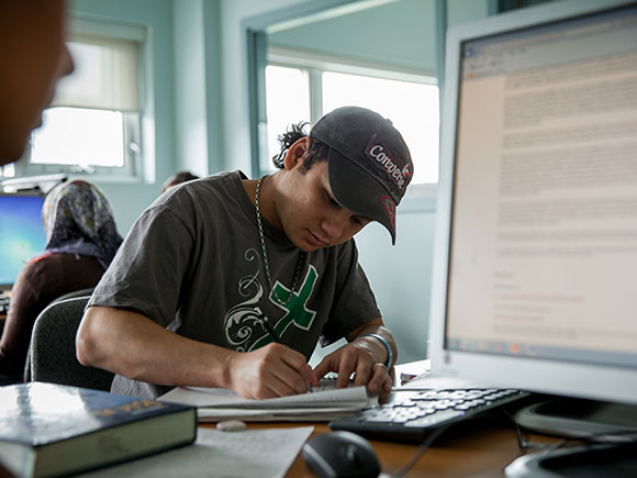 A student sits at a table in a classroom and writes notes. Another student uses a computer in the background.