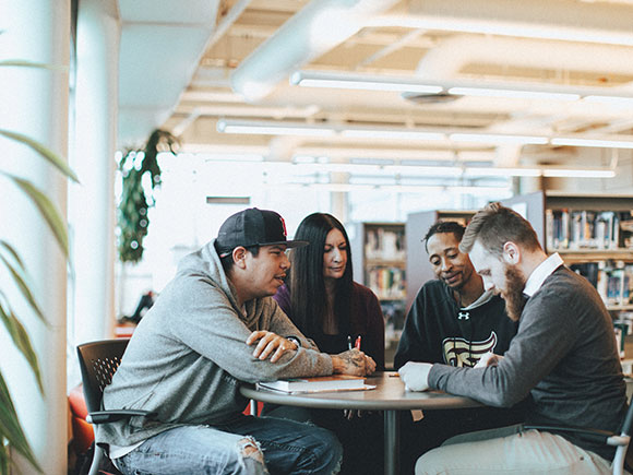 A group of four students study together with books open at a round table in a library.