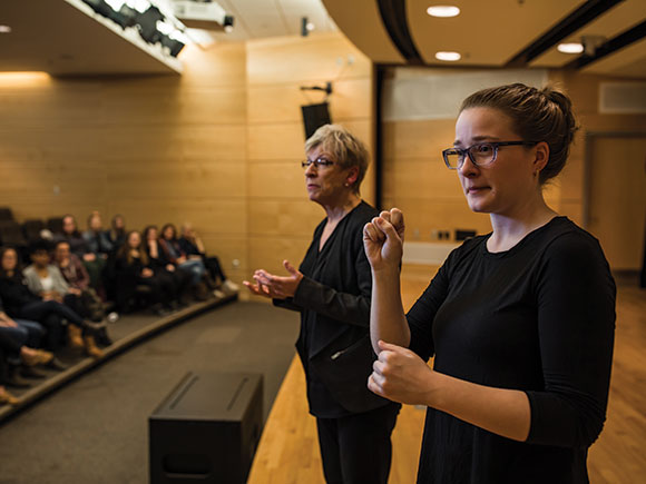 Two people wearing black practise ASL interpretation on a stage in front of a seated audience.