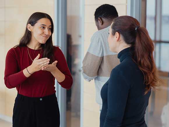 Two ASL students can be seen signing to one another outside their classroom while a third student attempts to enter the class