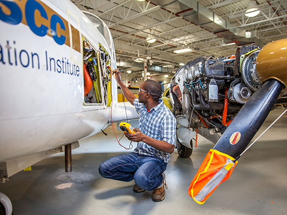 A student squats beside the body of an airplane. Part of the side is removed to display an electrical panel and he is connect