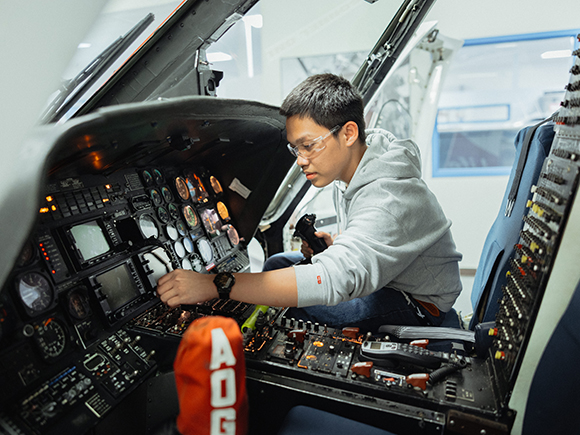 A student wearing safety glasses works on the inside electronics panel of an aircraft.
