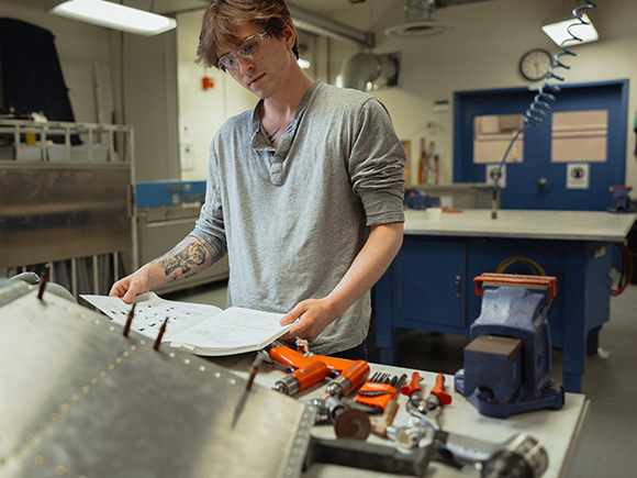 A student wearing safety goggles and a white t-shirt works on an aircraft component.