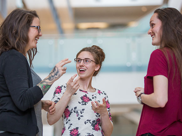 Three smiling people stand in college setting while communicating in American Sign Language.