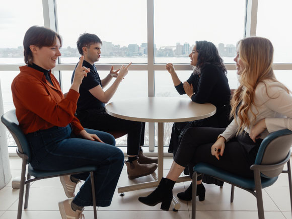 Three women stand against a blurred background and speak to each other in ASL.