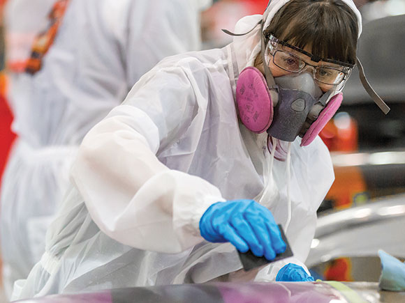 A woman in white coveralls, blue latex gloves, safety goggles and an industrial respirator sands down a surface.