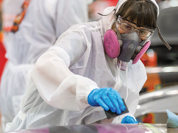 A student in coveralls, latex gloves, safety goggles and an industrial respirator sands down a surface.