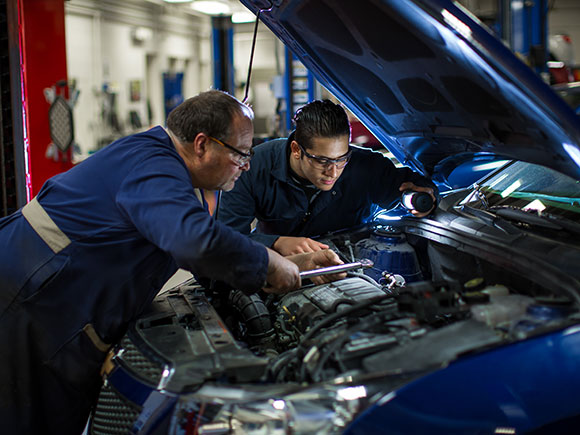 Two students work under the open hood of a car in an automotive workshop.