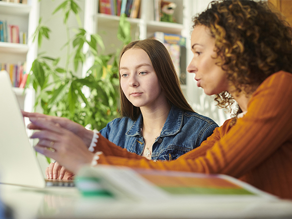 Two women sit at a table and look at a computer screen in a brightly lit room.