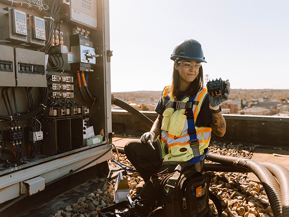 A woman in a safety vest and hard hat kneels looking at a piece of HVAC equipment.