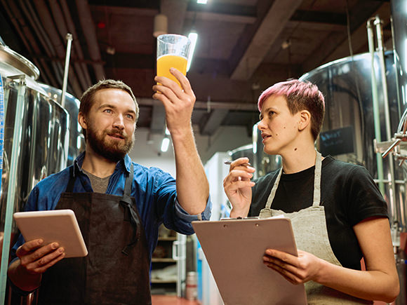 Two people wearing aprons stand in a brewery and look at a glass of beer while holding it up to the light. One carries a clip