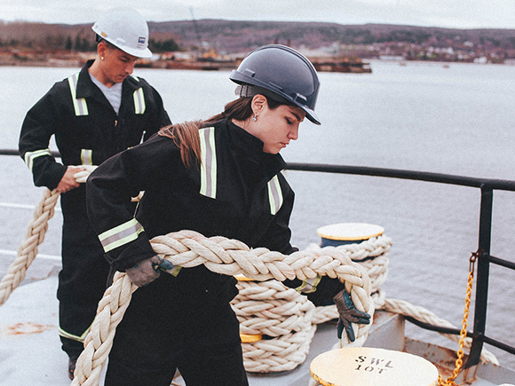 A man and a woman wearing coveralls and hardhats hold large ropes on the deck of a ship.