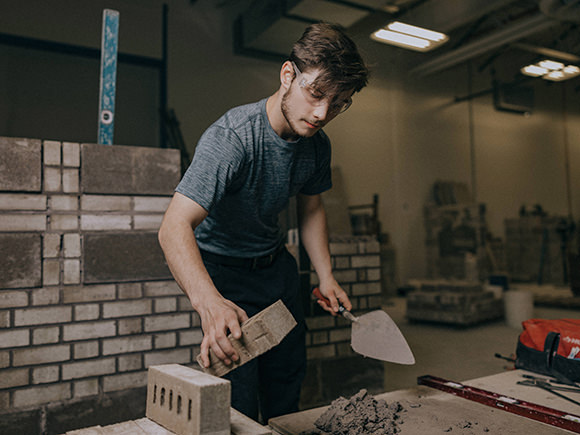 A student wearing safety glasses carefully places a brick in place at a work site.