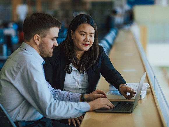 Two students look at an open laptop together. One of them points at the screen.