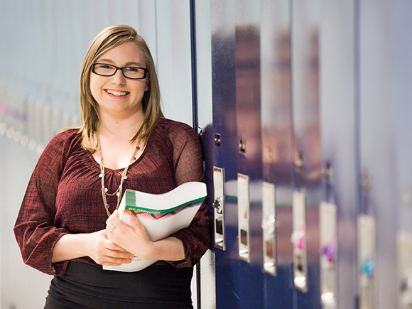 A student wearing a blouse, skirt, necklace and glasses stands in a hallway and leans against lockers while holding a book an