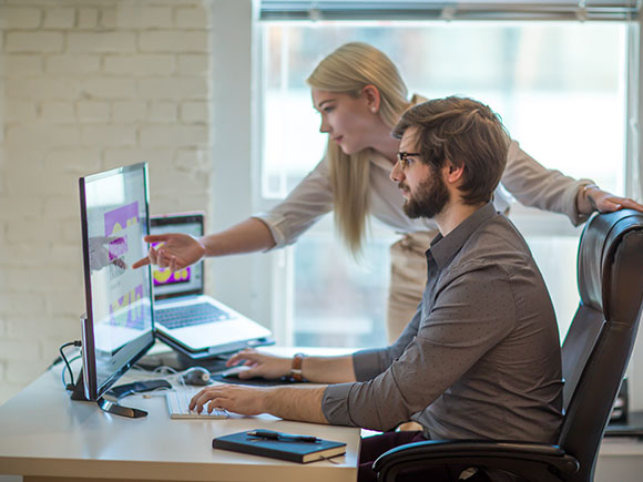 Two people look at a computer monitor together. One sits and types while the other stands and points at the screen.