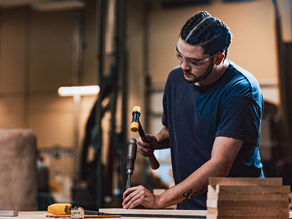 A student in a t-shirt and safety glasses uses a hammer and chisel in a campus woodshop.