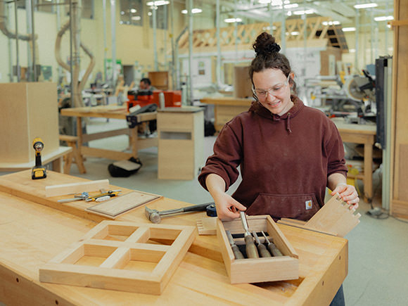 A student in a t-shirt and safety glasses uses a hammer and chisel in a campus woodshop.