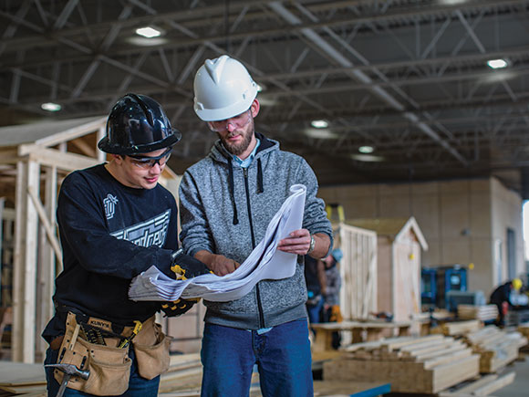 Two carpentry students wearing hard hats and safety glasses look at printed construction plans.