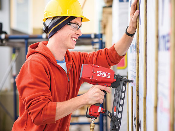 A carpentry student wearing a sweater, safety glasses and a hard hat uses a tool to construct a wooden frame for a wall.