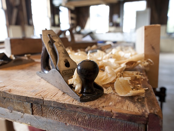 A hand plane tool used for restoration carpentry sits on a wooden table with wood shavings piled near by.