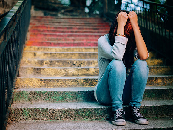A teenage girl in blue jeans and a grey sweatshirt sits on an outdoor staircase with her head in her hands.