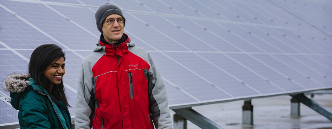 Smiling man and woman in winter clothing standing side by side in front of a large outdoor solar panel array.