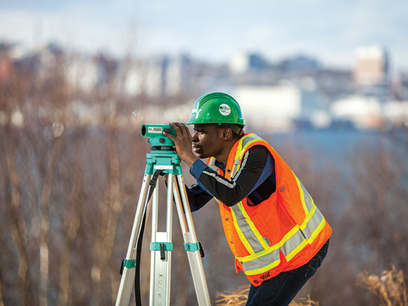A student in a safety vest and hard hat bends and looks through a piece of surveying equipment.