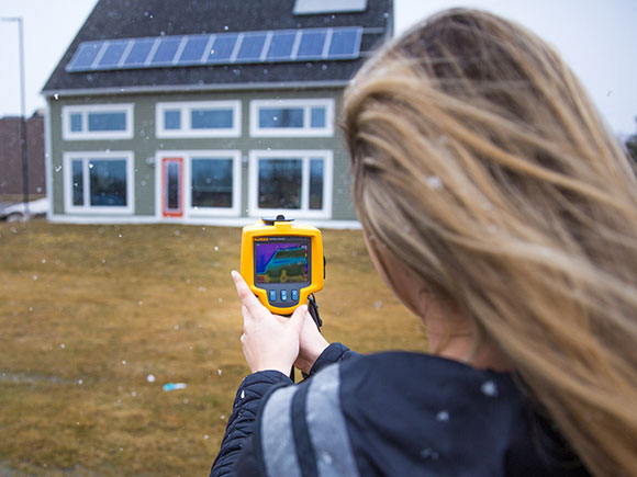 A person stands outdoors in front of a home and looks at the screen of a heat imaging tool.