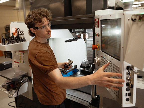 A student sits at a workbench inspecting a manufactured metal part. They are looking down, wearing safety glasses and hold