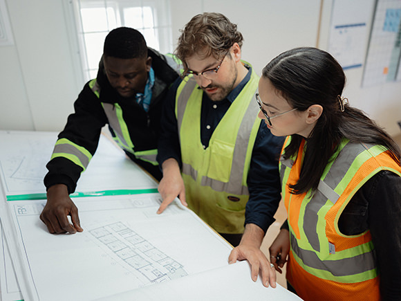 Two people in hard hats, safety glasses and safety vests look at a clipboard in a workshop.