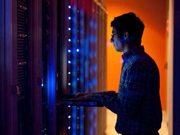 A man in a plaid shirt and glasses works in a computer server room.